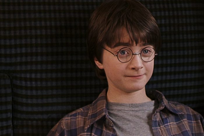 A young boy wearing round glasses and a plaid shirt sits, smiling slightly, against a dark striped background.