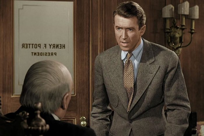 A man in a suit stands speaking to an elderly seated figure, in an office labeled 