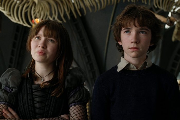 Two teenagers stand indoors, looking upward, framed by overhead skeletal structures in a dimly lit room. One wears gothic-style clothing, the other casual.