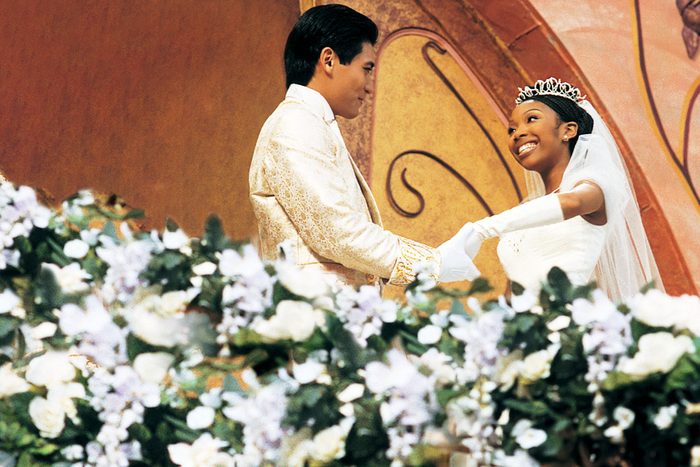 A couple smiles at each other, holding hands on a stage adorned with floral decorations and elaborate golden decor.