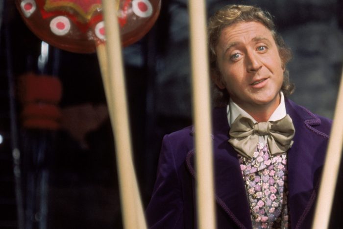 A person with curly hair smiles, wearing a purple coat and bow tie, standing behind striped candy sticks in a candy factory.