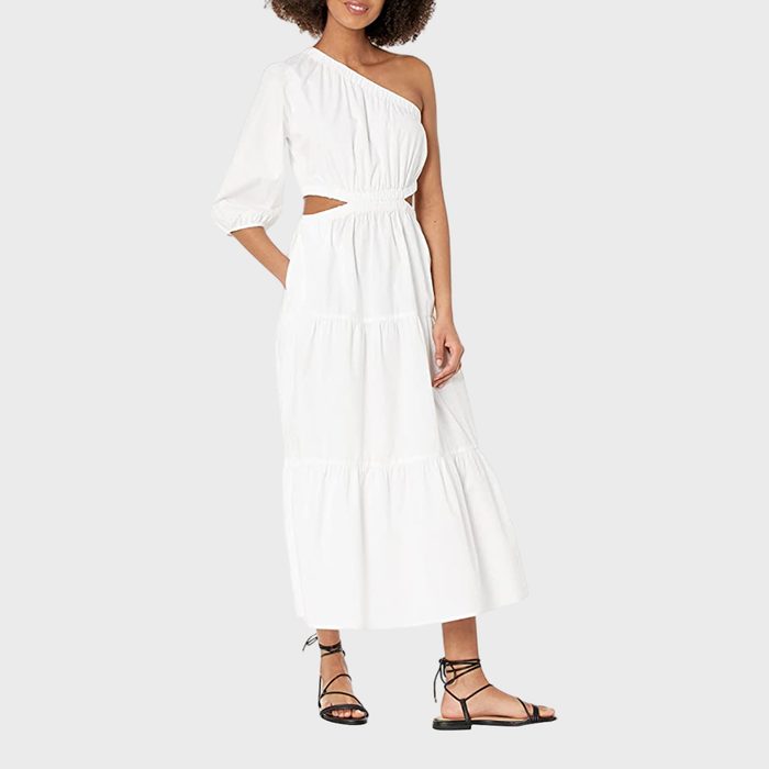 A woman wears a white, one-shoulder, cutout dress with black strappy sandals, standing against a plain gray background.