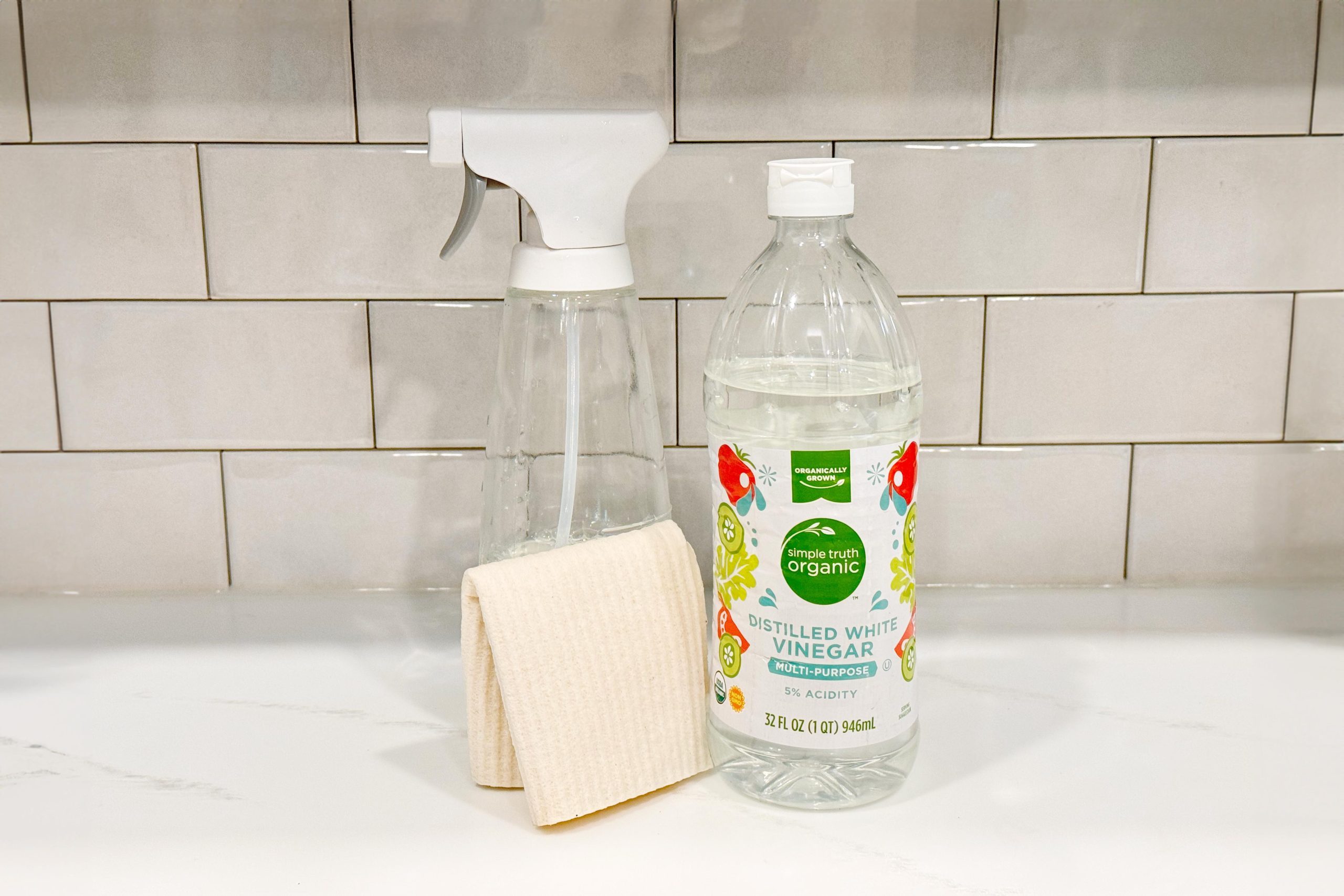 Spray bottle and vinegar bottle are positioned on a countertop, accompanied by a folded cloth, against a tiled backsplash.