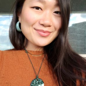 Woman smiling, wearing large earrings and a pendant necklace, with a glass window and blue sky in the background.