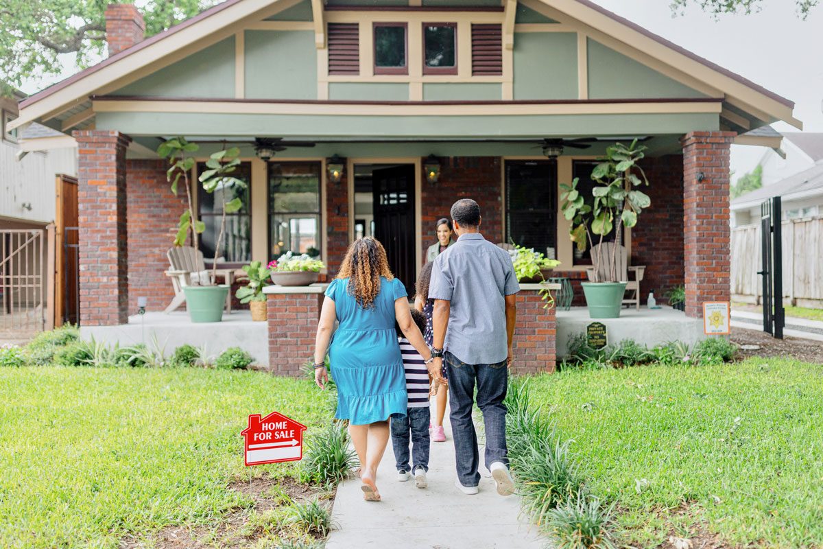 A couple walking into a house up for sale in an upscale and lush neighbourhood with a realtor standing in front of the house.